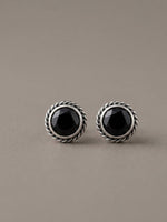 A pair of silver-toned round stud earrings with faceted black onyx centers, set in a textured rope-style bezel, on a neutral taupe background.