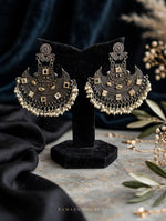 A pair of ornate silver-toned huggie earrings with intricate filigree, square gold-toned accents, and dangling pearl-like beads, displayed on a black velvet stand. The background features draped black fabric, a textured gray surface, and decorative olive branches.