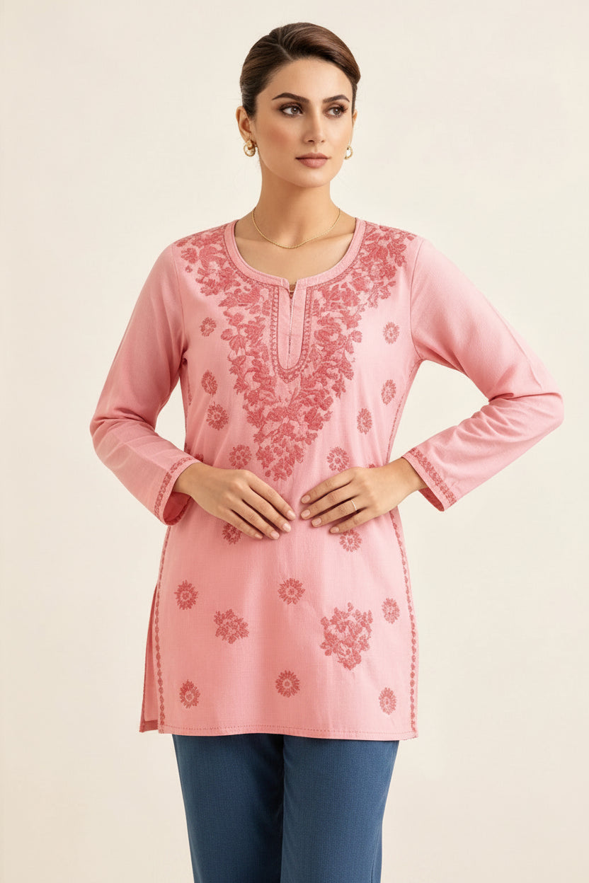 Front view of a model wearing a blush pink cotton short kurti featuring intricate pink floral embroidery, a keyhole neckline, and a square hem line, paired with matching trousers and her hands resting gently at her waist.