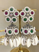 Boho-glam silver-finish jhumka earrings featuring white pearl clusters, green and maroon gemstones, and hanging white cowrie shells displayed on a gold stand.