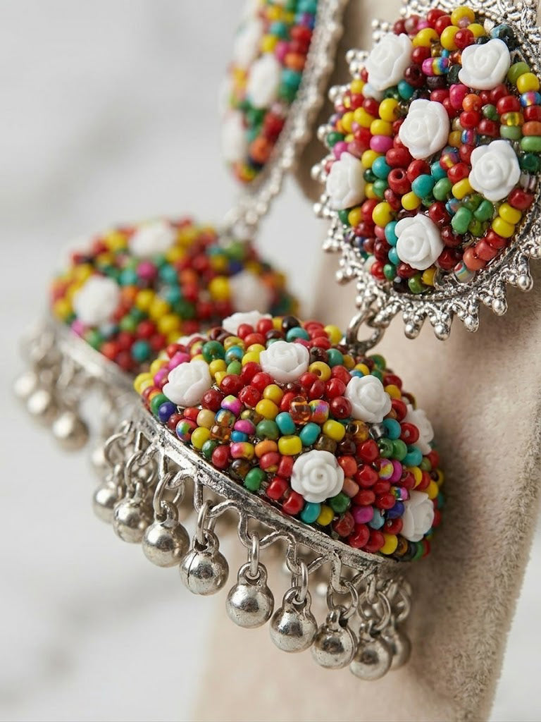 A pair of traditional Indian jhumka earrings displayed on a beige jewelry stand. The earrings feature silver-tone metal frames adorned with multicolored seed beads, white rose-shaped embellishments, and dangling silver bell charms.