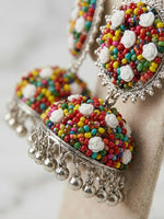 A pair of traditional Indian jhumka earrings displayed on a beige jewelry stand. The earrings feature silver-tone metal frames adorned with multicolored seed beads, white rose-shaped embellishments, and dangling silver bell charms.