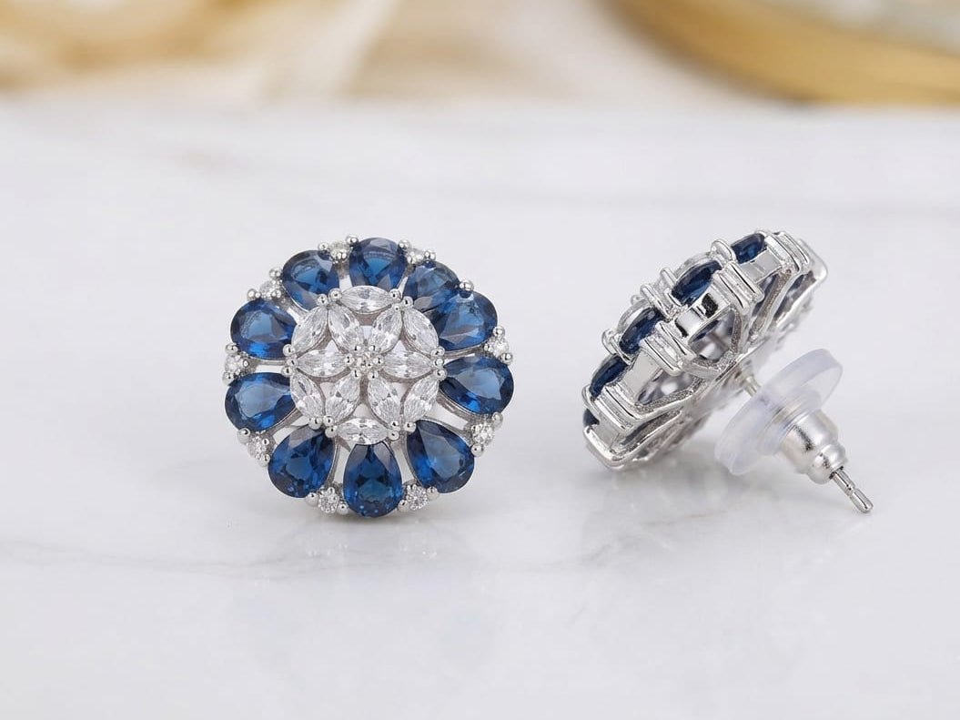 A product photo of a pair of silver floral cluster earrings featuring deep navy-blue pear-shaped stone petals surrounding a center cluster of sparkling clear marquise stones, with one earring shown from the front and the other angled to reveal the back post and setting.