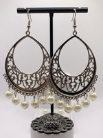 A pair of silver-toned filigree dangle earrings with white pearl-like drops, displayed on a black jewelry stand against a light gray background.