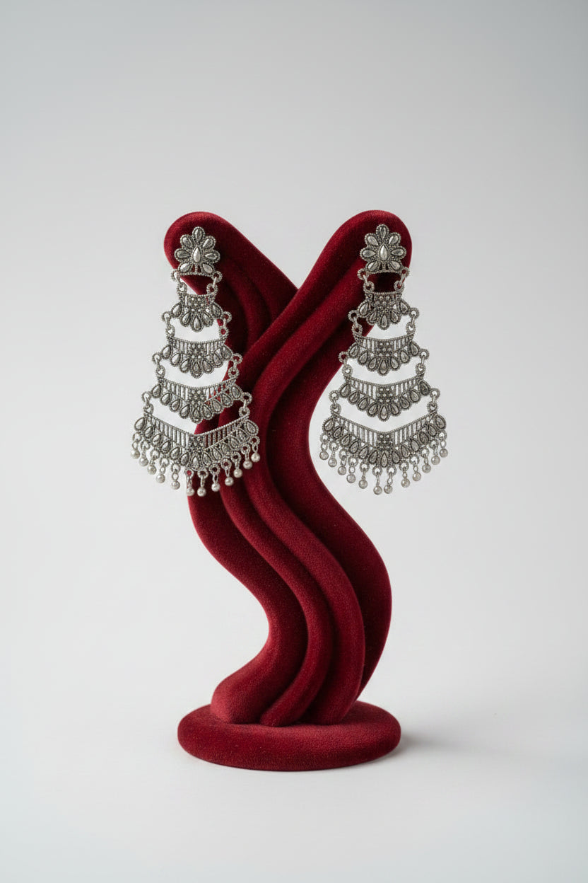 A pair of silver-toned, tiered dangle earrings displayed on a gold-colored wire stand with a dark wood base, set against a dark red curtain and white satin fabric.