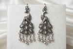 A close-up of a silver-toned traditional Indian jhumka earring worn on a person's ear. The ornate dangling earring features a peacock motif with intricate filigree detailing and hanging silver bells.