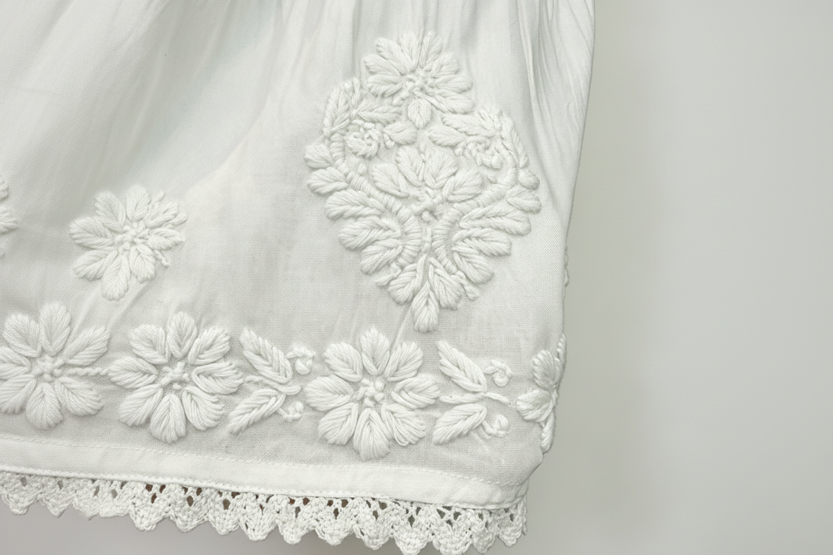 Close-up of a white embroidered fabric garment, showing floral embroidery and a scalloped lace hem against a white background.
