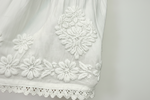 Close-up of a white embroidered fabric garment, showing floral embroidery and a scalloped lace hem against a white background.