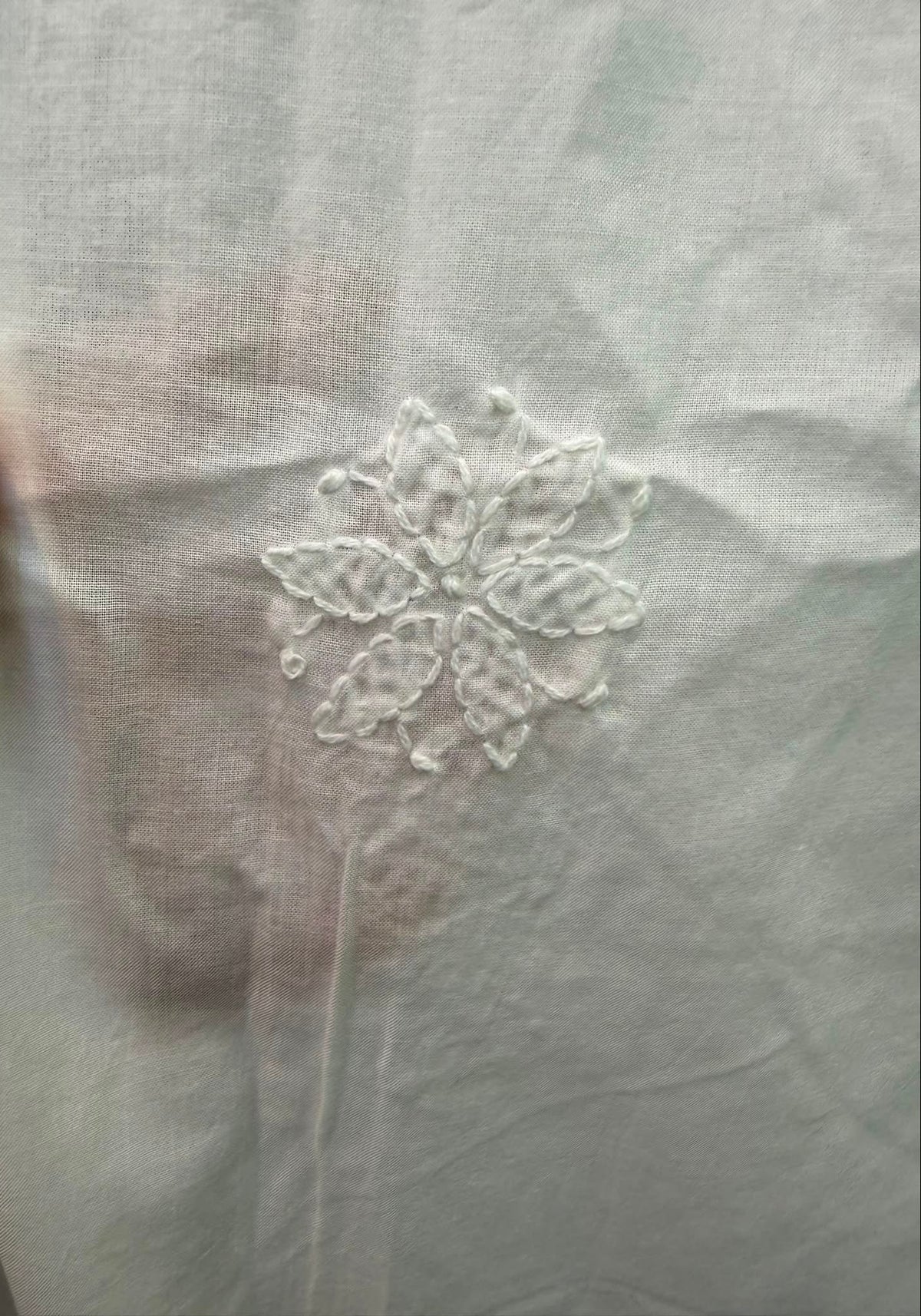 Close-up of a white fabric surface with a small white embroidered floral motif, with a finger visible at the left edge holding the fabric.