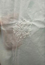 Close-up of a white fabric surface with a small white embroidered floral motif, with a finger visible at the left edge holding the fabric.