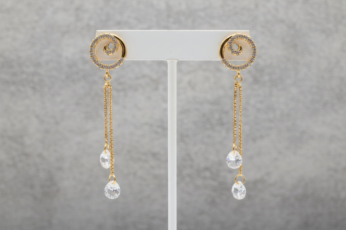  Close-up of the Vortex rose gold polish earrings on a display stand, highlighting the spiral stone setting and the dual-chain white stone drops.
