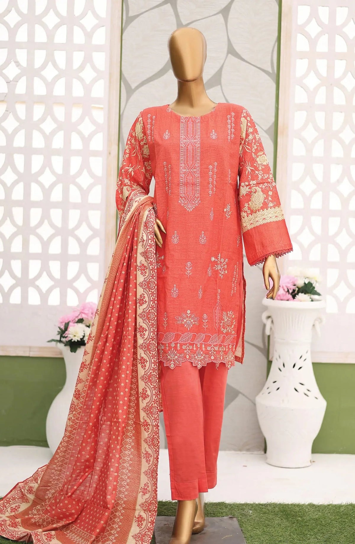 Coral pink traditional outfit with embroidery on a mannequin against a decorative white wall.