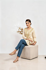 Full-length seated view of a model wearing a creamy beige cotton short kurti with gold embroidery, paired with matching trousers, a square hem line, and holding a gold clutch while sitting on a block seat.