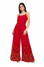 A close-up studio photograph focusing on the lower half of a woman wearing flowing, wide-leg red palazzo pants. The sheer fabric pants feature intricate, multi-colored floral and vine embroidery in shades of gold, orange, and green along the bottom hem and lower leg section. The upper part of her body and the top she is wearing are cropped out of the frame, making the embroidered palazzo pants the central focus. She is standing against a plain white background, and the lighting is even and bright.