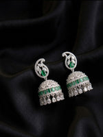 A pair of silver-toned Indian jhumka earrings with green emerald-cut stones, displayed on a draped black satin fabric background.