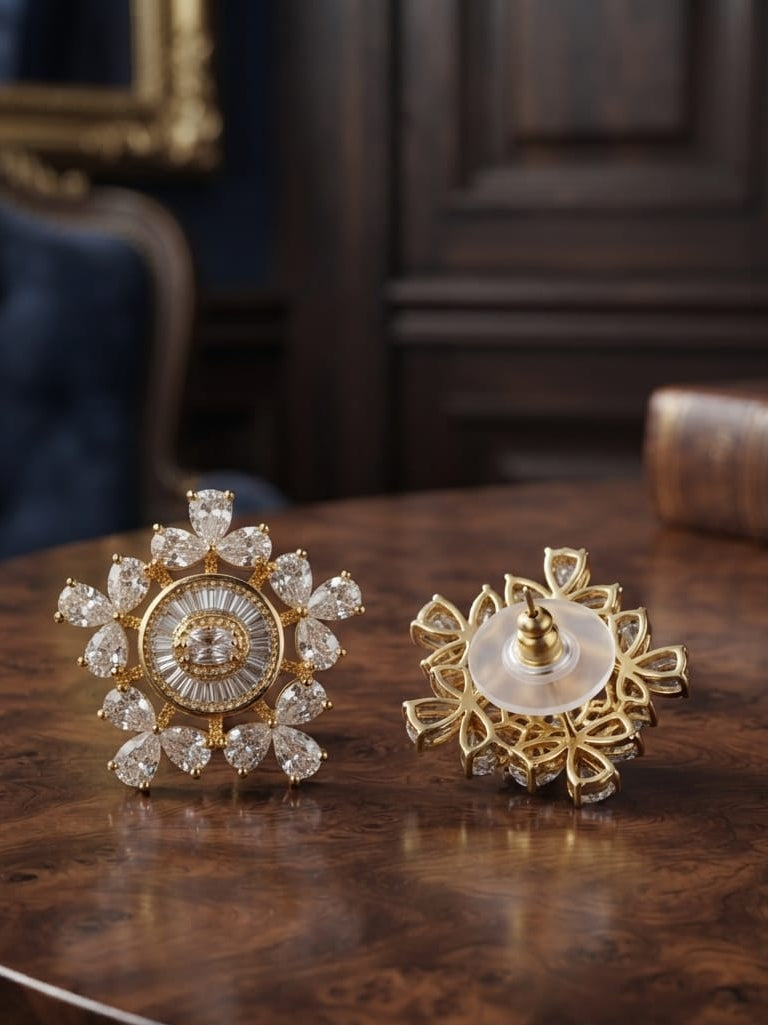 Two ornate gold-toned brooches displayed on a polished wooden table in an elegant interior setting. One brooch is adorned with clear crystals arranged in a floral or snowflake-like pattern, while the other shows the back with a circular white stone and gold filigree detailing.