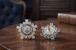A pair of silver-toned snowflake-shaped brooches with clear gemstones, displayed on a polished dark wood table. One brooch is shown from the front, revealing a detailed sunburst design with a central oval motif.