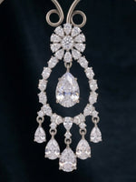 An ornate silver-toned chandelier drop earring featuring a large pear-cut clear center stone surrounded by smaller round and marquise-cut clear stones, with five dangling teardrop stones below, set against a dark navy background.