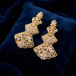 Close-up of elegant gold-toned statement earrings with detailed filigree patterns and marquise-cut crystals, resting on a luxurious dark blue velvet cushion.