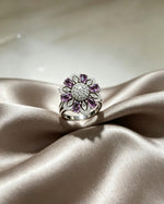 Silver-tone floral halo ring with deep purple oval stones and sparkling AD center, displayed on champagne satin fabric.