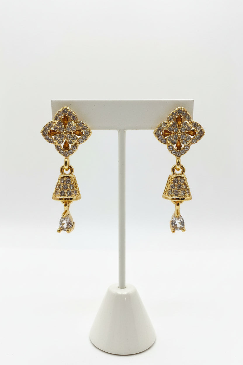 Detailed view of golden brass earrings featuring a shimmering pave-set starburst stud and a dainty, crystal-encrusted bell-shaped drop hanging from a delicate link.