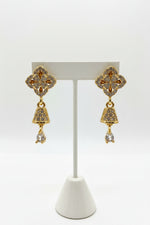 Detailed view of golden brass earrings featuring a shimmering pave-set starburst stud and a dainty, crystal-encrusted bell-shaped drop hanging from a delicate link.