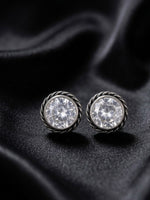 A pair of silver-toned round stud earrings with clear faceted gemstones, displayed on a draped black satin fabric background.