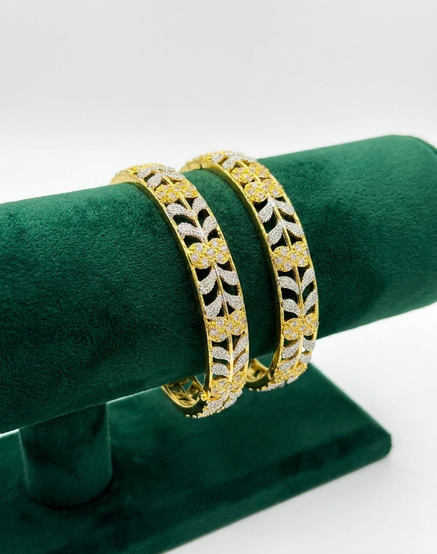 Close-up shot of a pair of dual-tone American Diamond bangles featuring an intricate gold floral lattice and silver leaf vine pattern, displayed on a green velvet stand