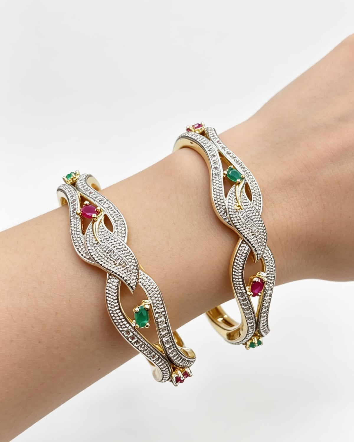 Close-up of a person's wrist wearing two dual-tone floral lattice bangles, featuring a silver-toned pavé finish with white American Diamonds and contrasting ruby and emerald gemstone accents.
