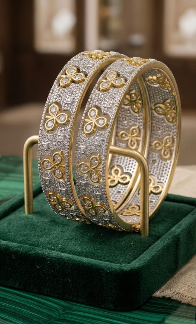 Close-up shot of a pair of dual-tone luxury bangles featuring gold-plated triple-petal floral motifs encrusted on a shimmering silver pavé stone-set band, displayed on a green velvet stand.