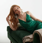 A model wearing elegant chandelier earrings with emerald green stones and cubic zirconia detailing while lying on a dark green velvet sofa.