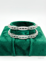 A pair of silver-toned American diamond bangles with a unique wavy, scalloped edge and intricate open-work lattice design, displayed on a plush emerald green velvet jewelry stand.