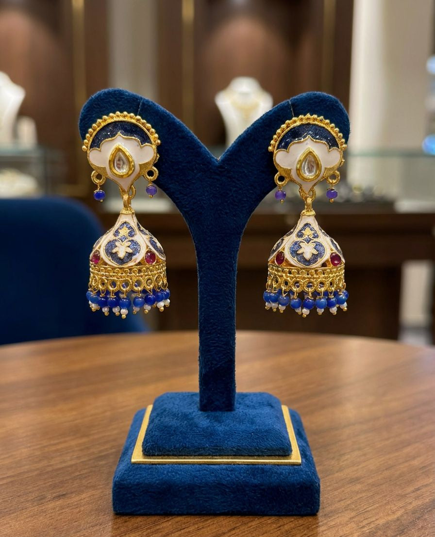 Elegant gold-plated brass jhumka earrings featuring intricate blue and white enamel Meenakari work, displayed on a blue stand.