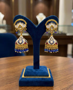 Elegant gold-plated brass jhumka earrings featuring intricate blue and white enamel Meenakari work, displayed on a blue stand.
