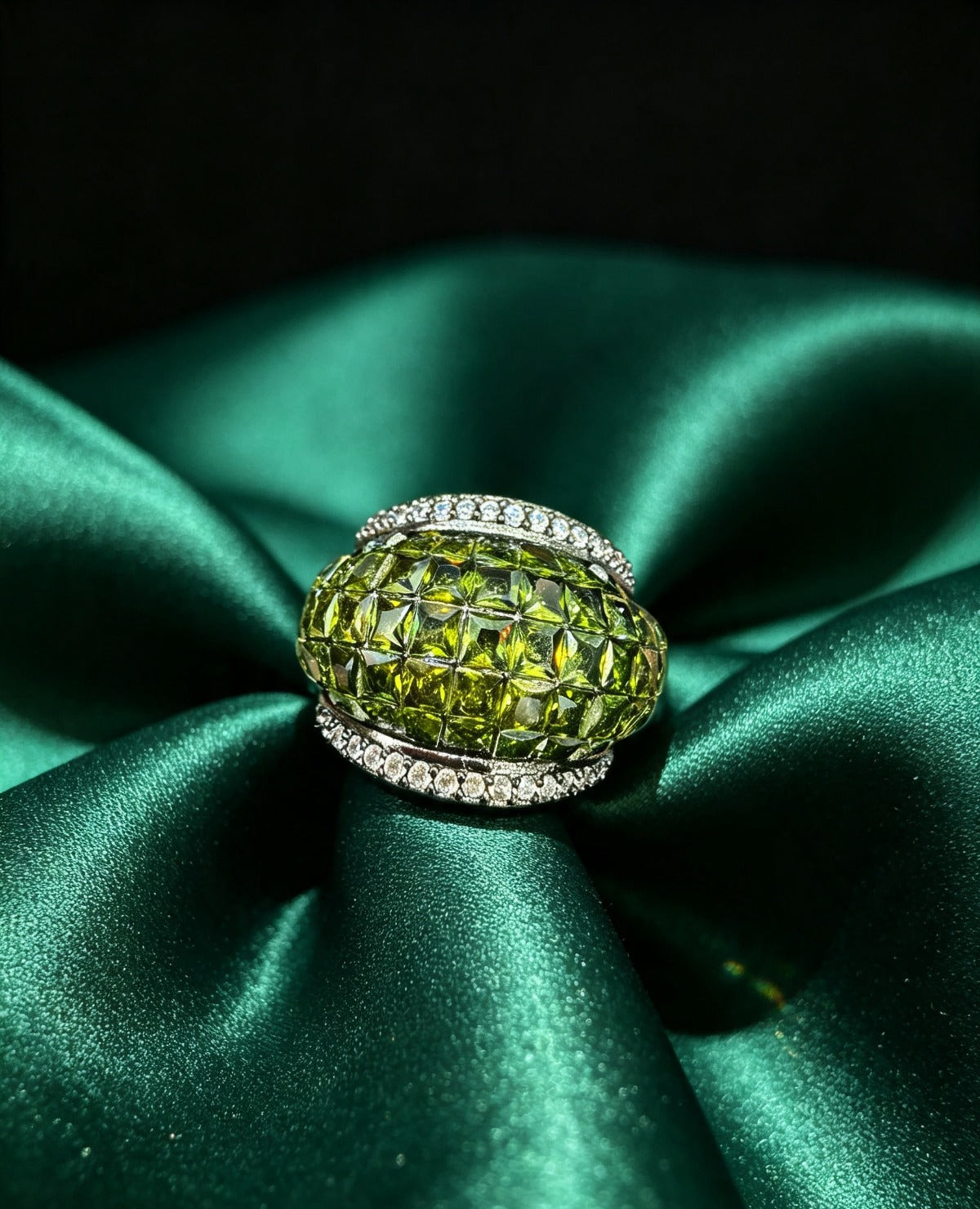 Elysia Green Dome Ring featuring a large faceted oval green stone with a checkerboard pattern, bordered by sparkling pave crystals, resting gracefully on a draped emerald satin fabric.