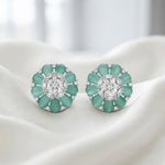 A pair of floral stud earrings with mint green oval stones and a central clear round stone, set in silver-toned metal and displayed on draped white fabric.