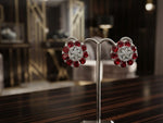 A pair of silver-toned earrings with red gemstone clusters and clear central stones, displayed on a metal stand on a dark wooden table with a blurred luxury interior in the background.