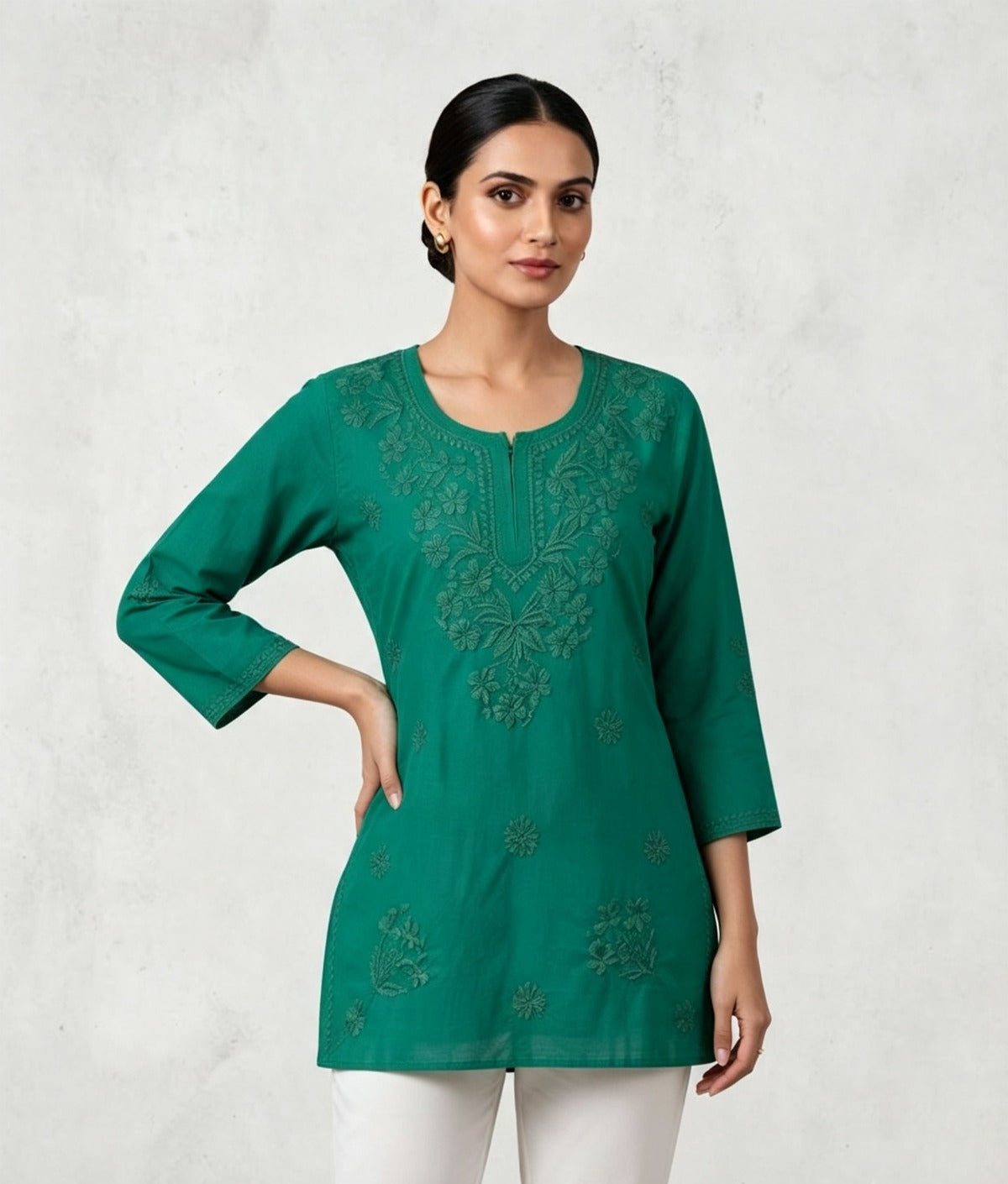 A woman modeling a vibrant green short kurti with her hand on her hip, featuring tonal embroidery along the split neckline and scattered floral motifs on the lower front, paired with white trousers.