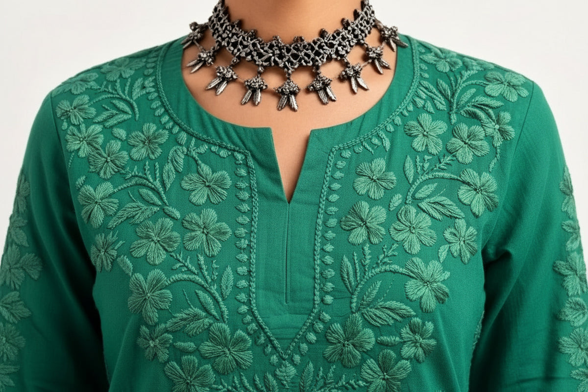 A close-up view of a person wearing a vibrant green short kurti with detailed tonal floral embroidery along the split round neckline, accessorized with an ornate silver-toned choker necklace.