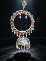 A large silver-toned traditional Indian earring featuring a circular frame adorned with alternating red and green square stones, a dangling bell-shaped charm with intricate filigree detailing, and small hanging silver beads, set against a dark background.