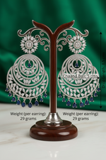 Statement 8.5cm silver Chaand Bali earrings featuring intricate clear crystal marquise patterns and a fringe of sapphire blue teardrop accent stones on a luxury product display stand