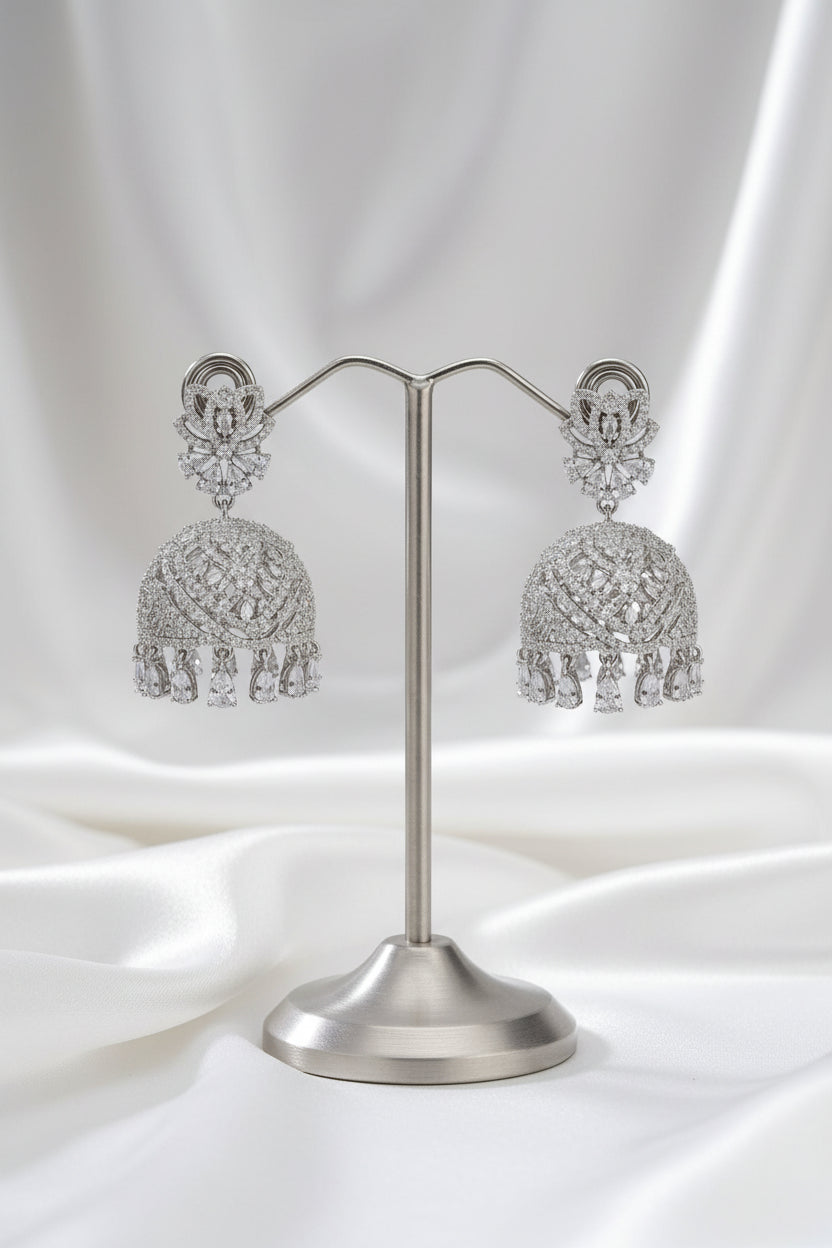 A pair of silver-toned Indian jhumka earrings with intricate filigree work and dangling tassel details, displayed on a silver stand against a black background.
