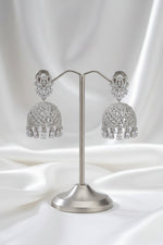 A pair of silver-toned Indian jhumka earrings with intricate filigree work and dangling tassel details, displayed on a silver stand against a black background.