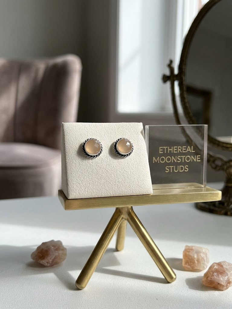 A pair of round moonstone stud earrings with silver-toned textured borders and peach-colored stones, displayed on a cream jewelry stand on a gold-colored mini table. A clear acrylic sign next to it reads 'ETHERAL MOONSTONE STUDS', and pink crystal points are scattered on the white surface in the foreground.