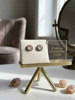 A pair of round moonstone stud earrings with silver-toned textured borders and peach-colored stones, displayed on a cream jewelry stand on a gold-colored mini table. A clear acrylic sign next to it reads 'ETHERAL MOONSTONE STUDS', and pink crystal points are scattered on the white surface in the foreground.