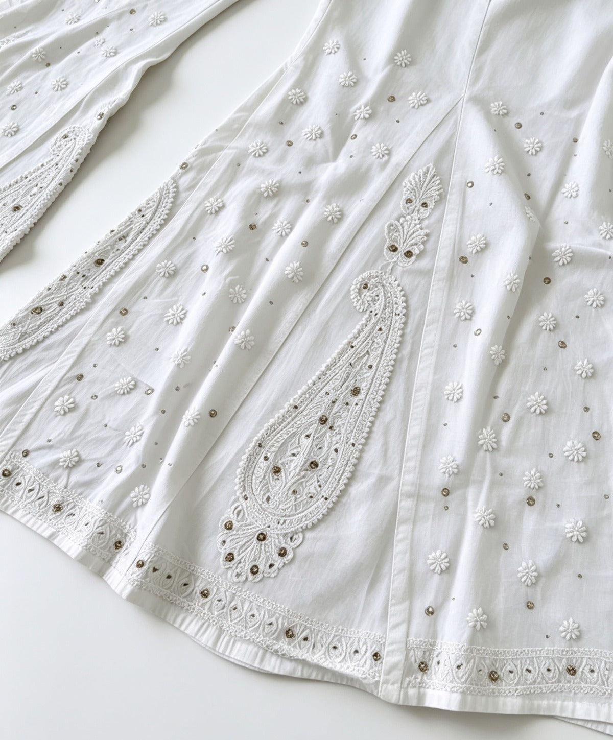 A flat-lay view of a pair of ethereal white sharara pants, showcasing delicate paisley motifs and intricate Bakhiya embroidery detail, set against a clean white background.
