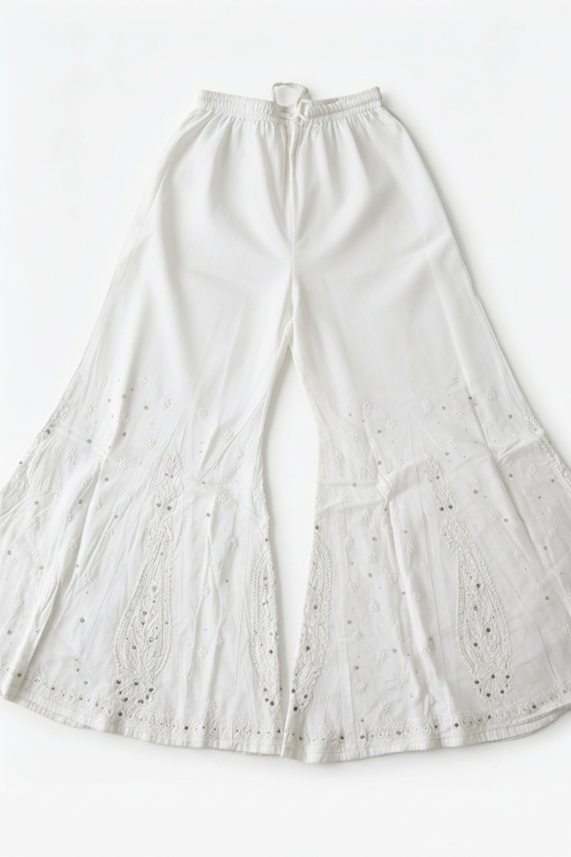 A flat-lay view of a pair of ethereal white sharara pants, showcasing delicate paisley motifs and intricate Bakhiya embroidery detail, set against a clean white background.