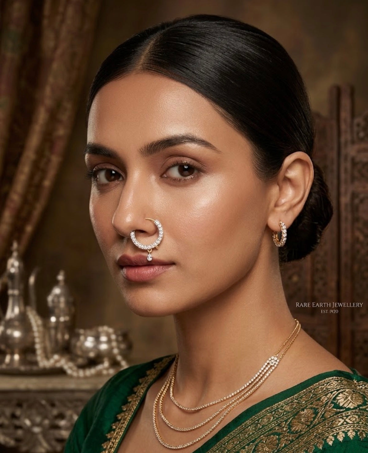 A close-up portrait of a woman wearing a silver crescent moon nose ring, matching hoop earrings, and layered gold and silver necklaces, paired with a green and gold embroidered sari. The background is a warm, traditional interior with ornate silver vessels.