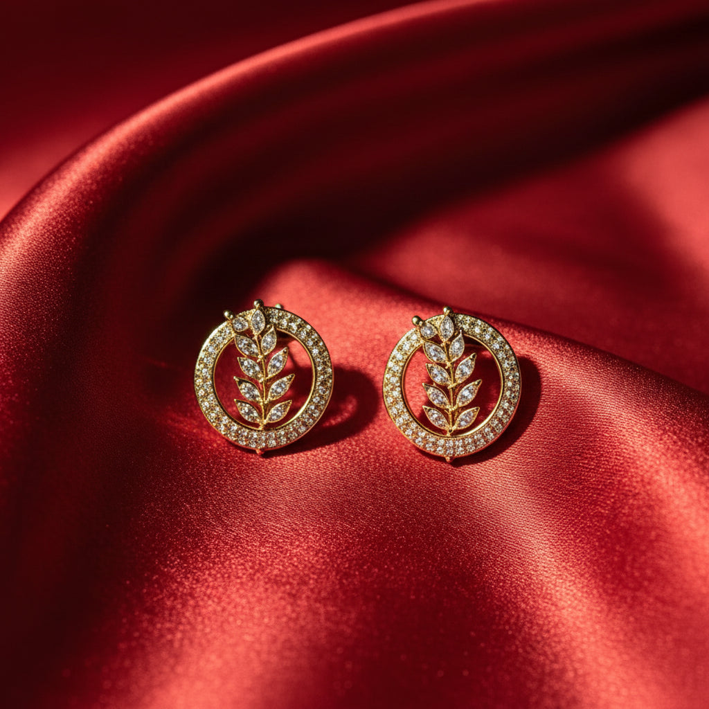 Famaza Couture Radiant Leaflet American Diamond Gold-Plated Studs – Pair of circular gold-toned earrings with sparkling AD leaf motif, beautifully displayed on luxurious emerald green silk fabric.