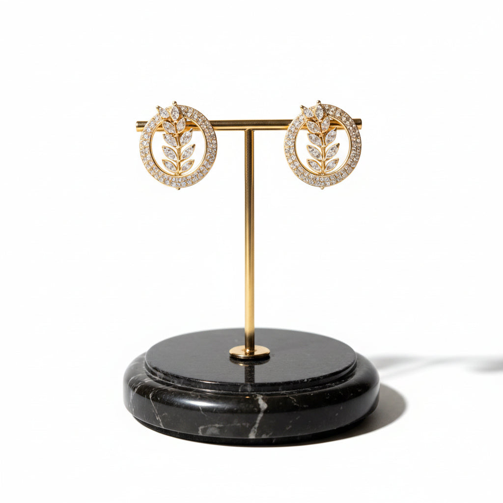 Famaza Couture Radiant Leaflet American Diamond Gold-Plated Studs – Pair of circular earrings with sparkling AD stones, central leaf motif, measuring 2 cm, displayed on elegant gold stand with black marble base.